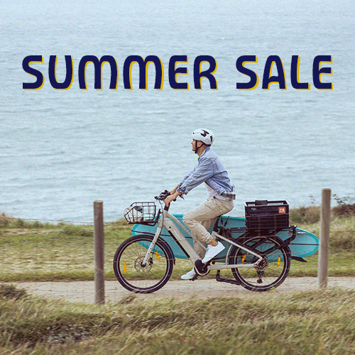 Summer Sale Monty Bikes : personne roulant à vélo cargo électrique en bord de mer avec une planche de paddle.